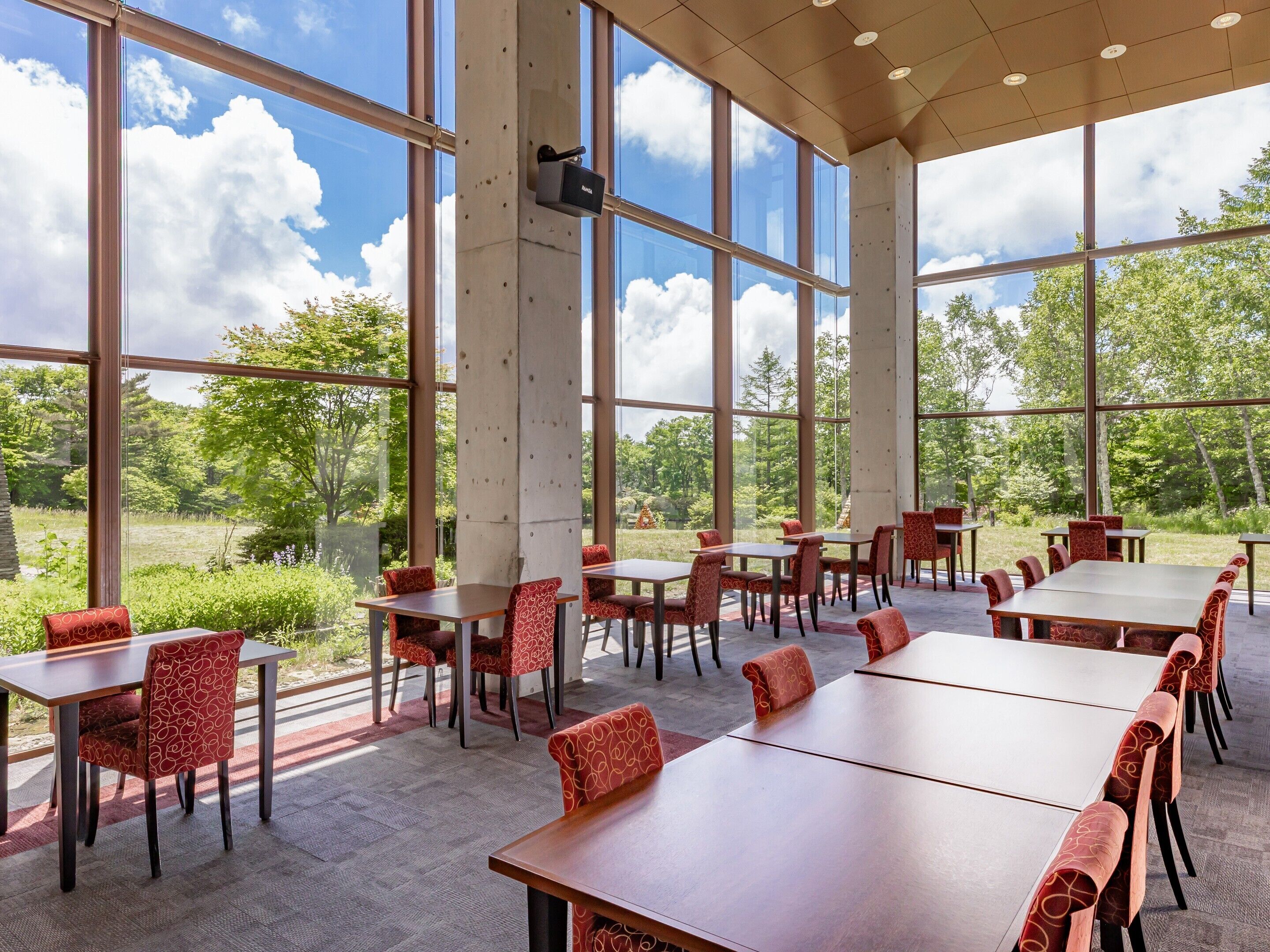 A spacious indoor area with large windows, multiple tables and chairs, and a view of greenery outside.