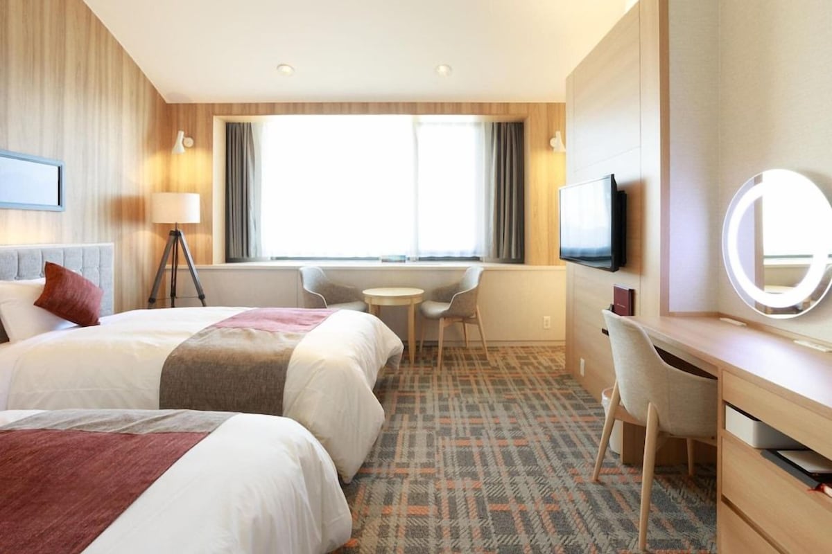 A hotel room with two beds, a desk, a chair, a TV, and a window with curtains.