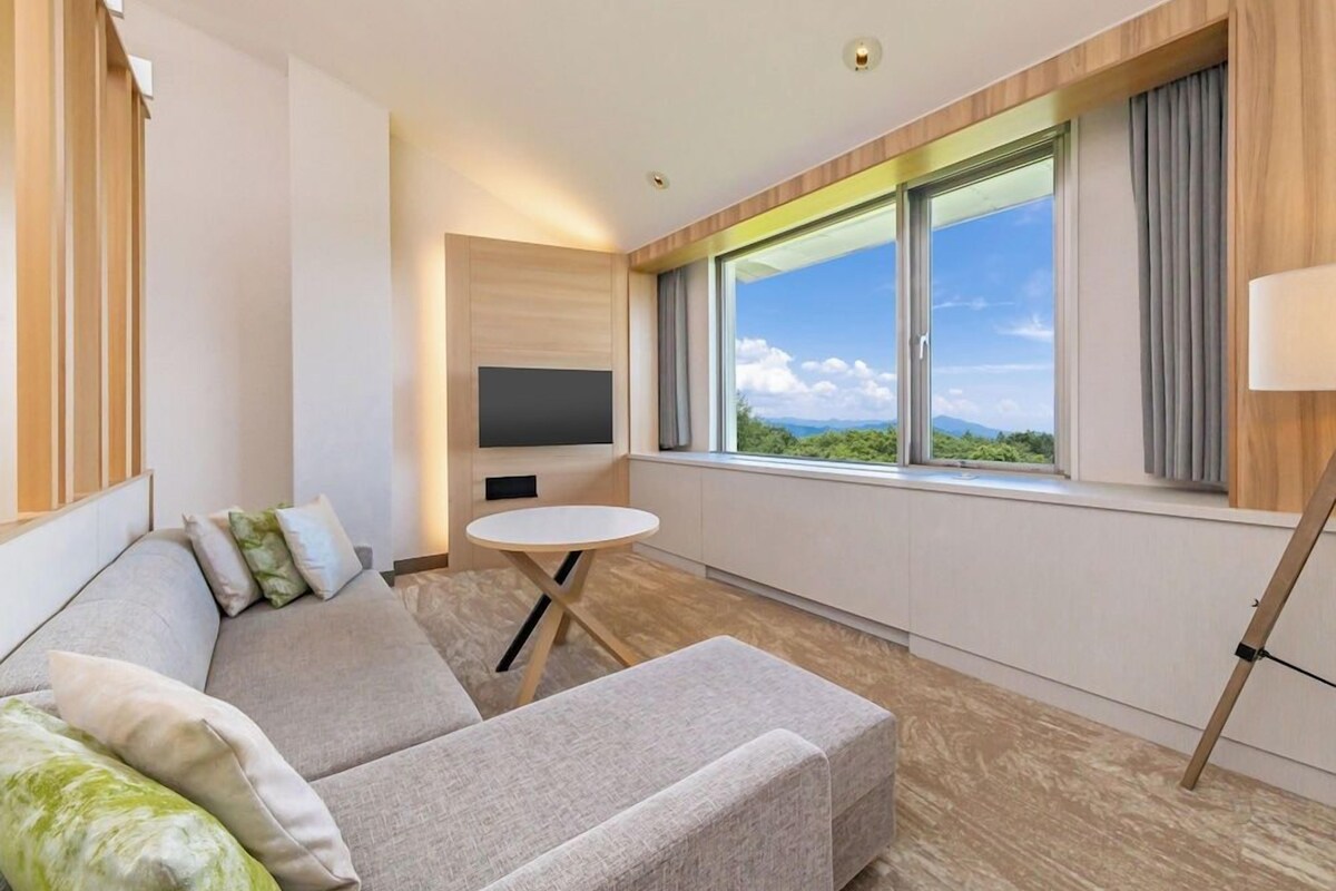 Et moderne hotelværelse med sofa, et rundt bord, fjernsyn og et vindue med udsigt over landskabet.