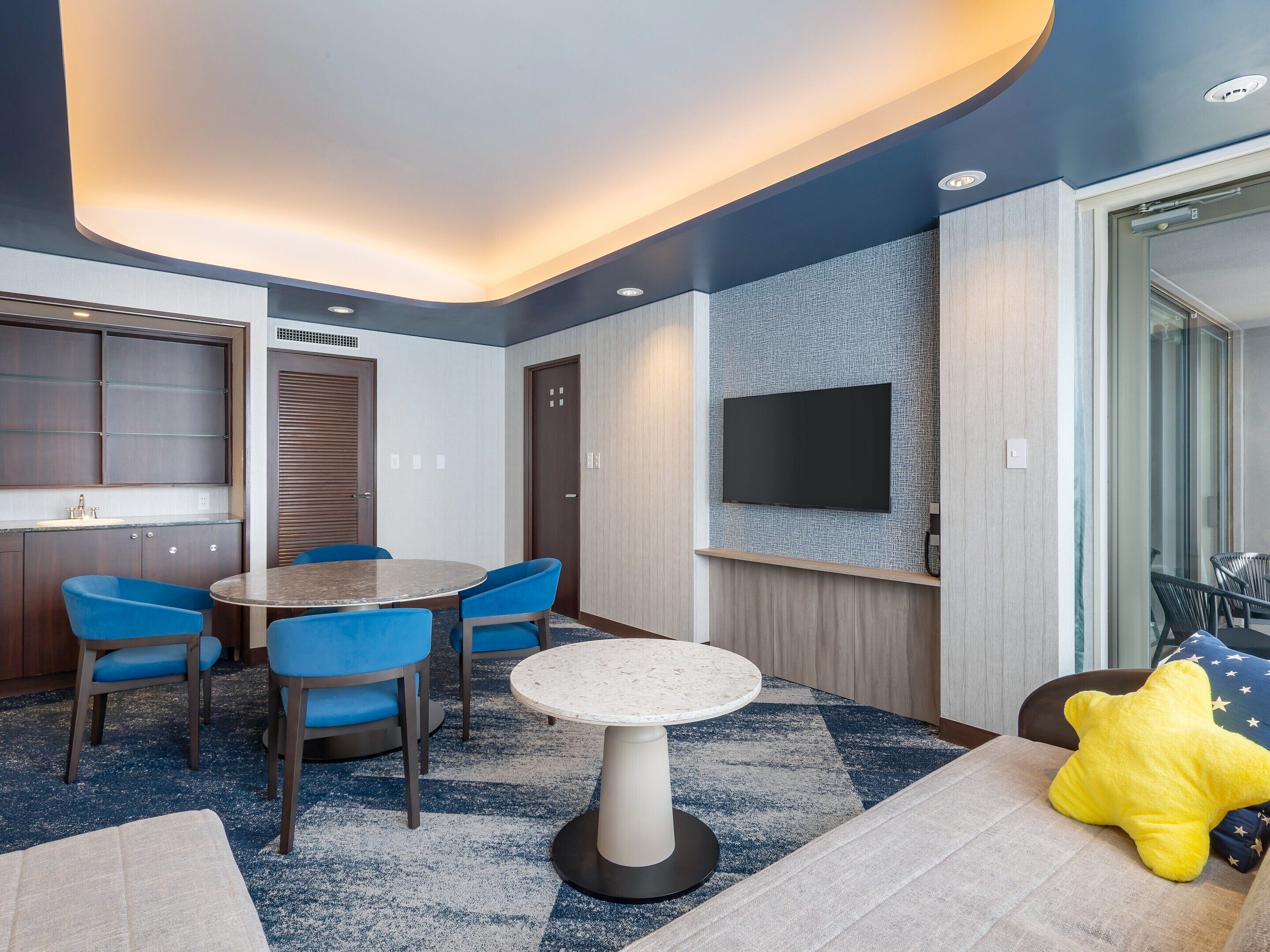 Una habitación de hotel moderna con una mesa de comedor redonda, sillas azules, un televisor de pantalla plana montado en la pared y un cojín con forma de estrella en el sofá.