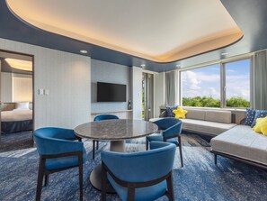 A modern hotel room with a round table, blue chairs, a sofa with star-patterned pillows, a flat-screen TV, and a large window with a view of greenery.
