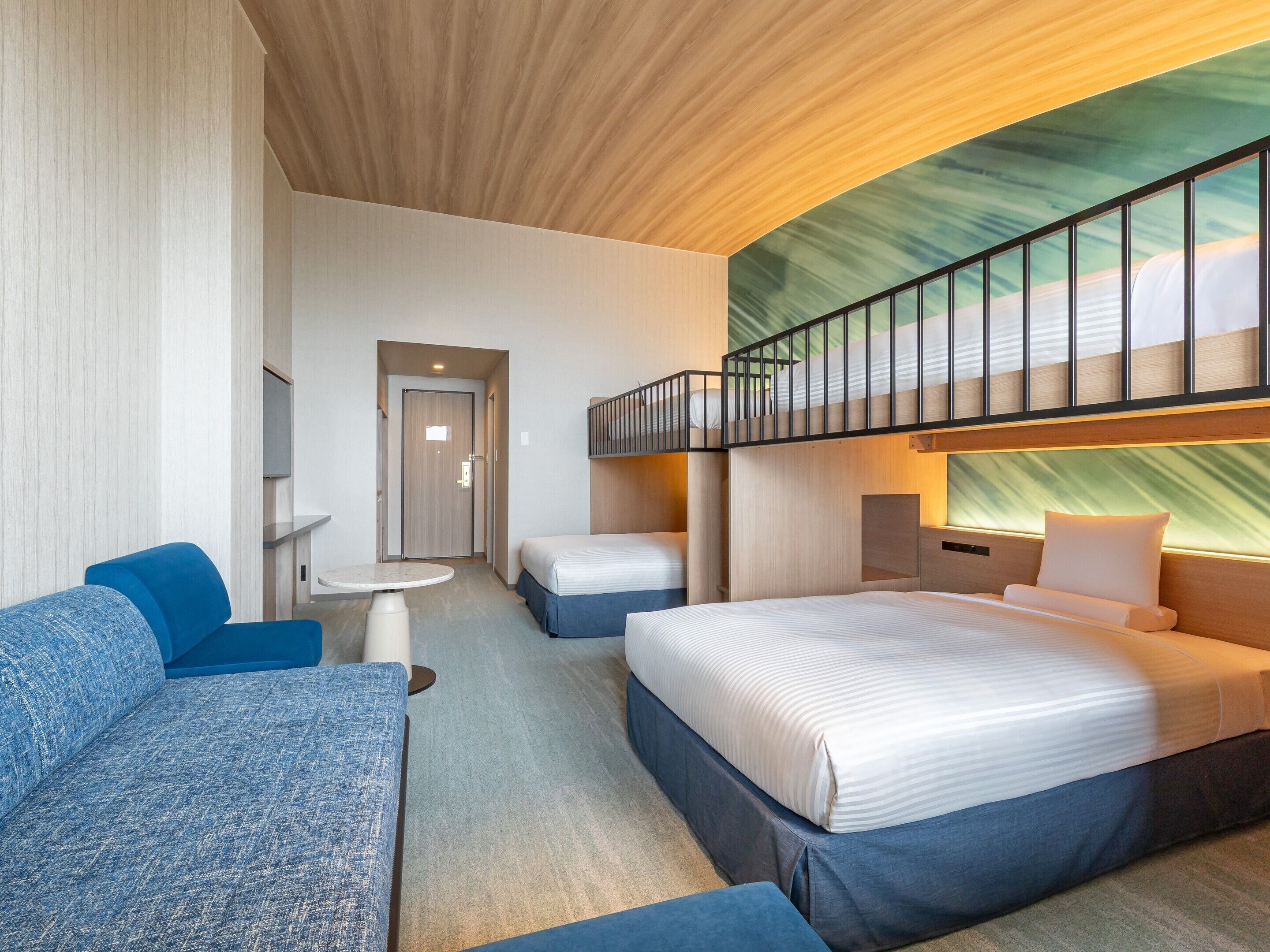 A modern hotel room with a bunk bed, a sofa, a small table, and a view of the sky through a large window.