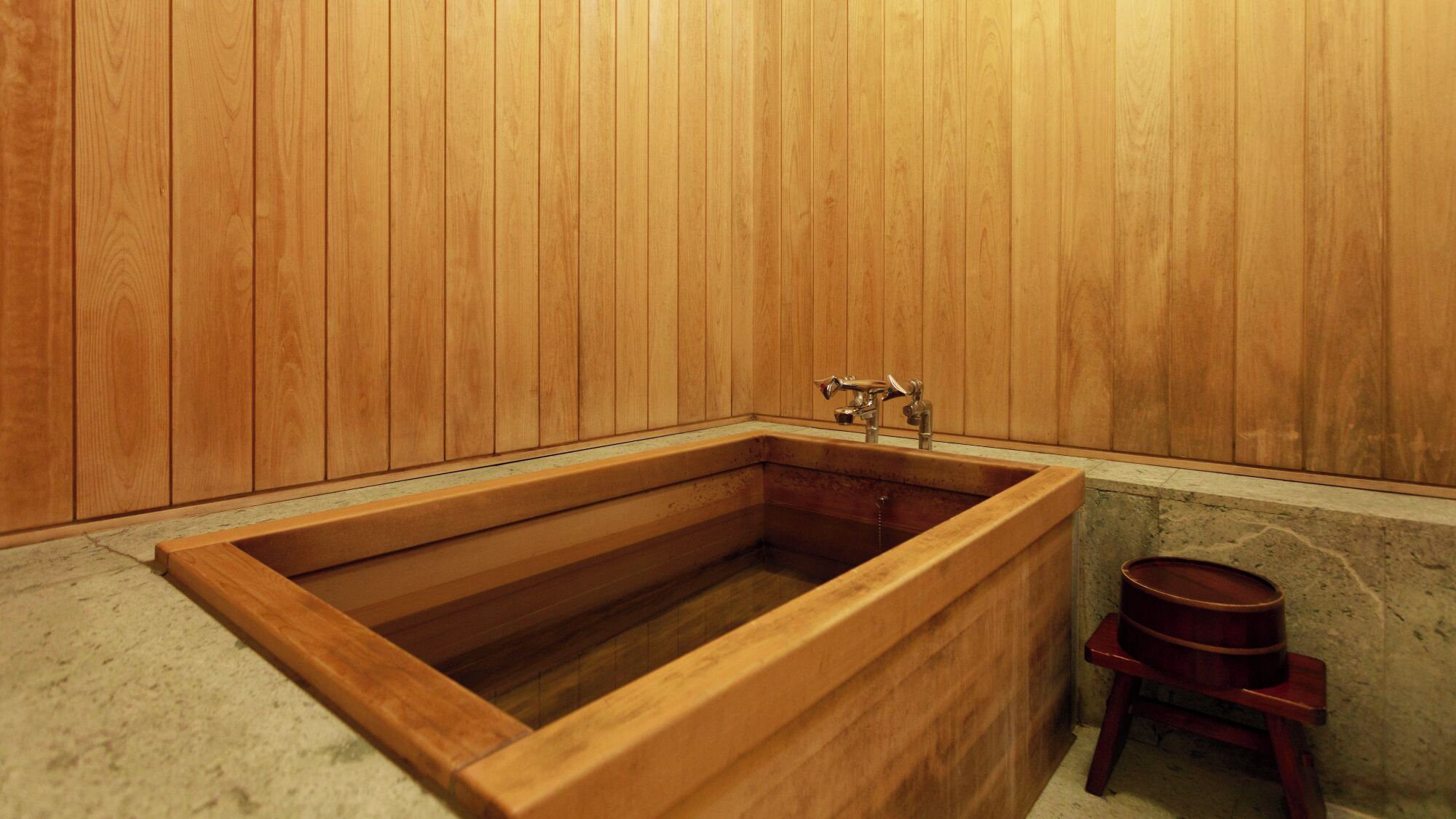 A wooden bathtub with a faucet, a wooden bench, and a stone wall.