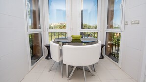 Habitación de esquina con una mesa redonda de vidrio y sillas blancas, ventanas amplias y vistas a zonas verdes y edificios.
