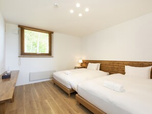 Una habitación de hotel moderna con dos camas individuales, un cabecero de madera, una mesita de noche con lámpara y una ventana con persianas.