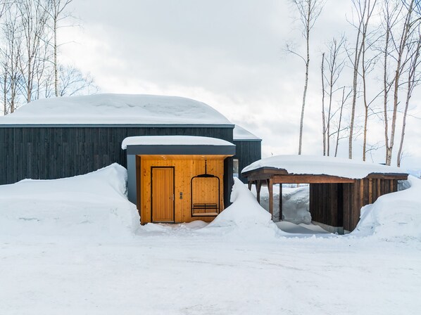 Una cabaña de madera con puerta y un pequeño porche, rodeada de nieve y árboles.