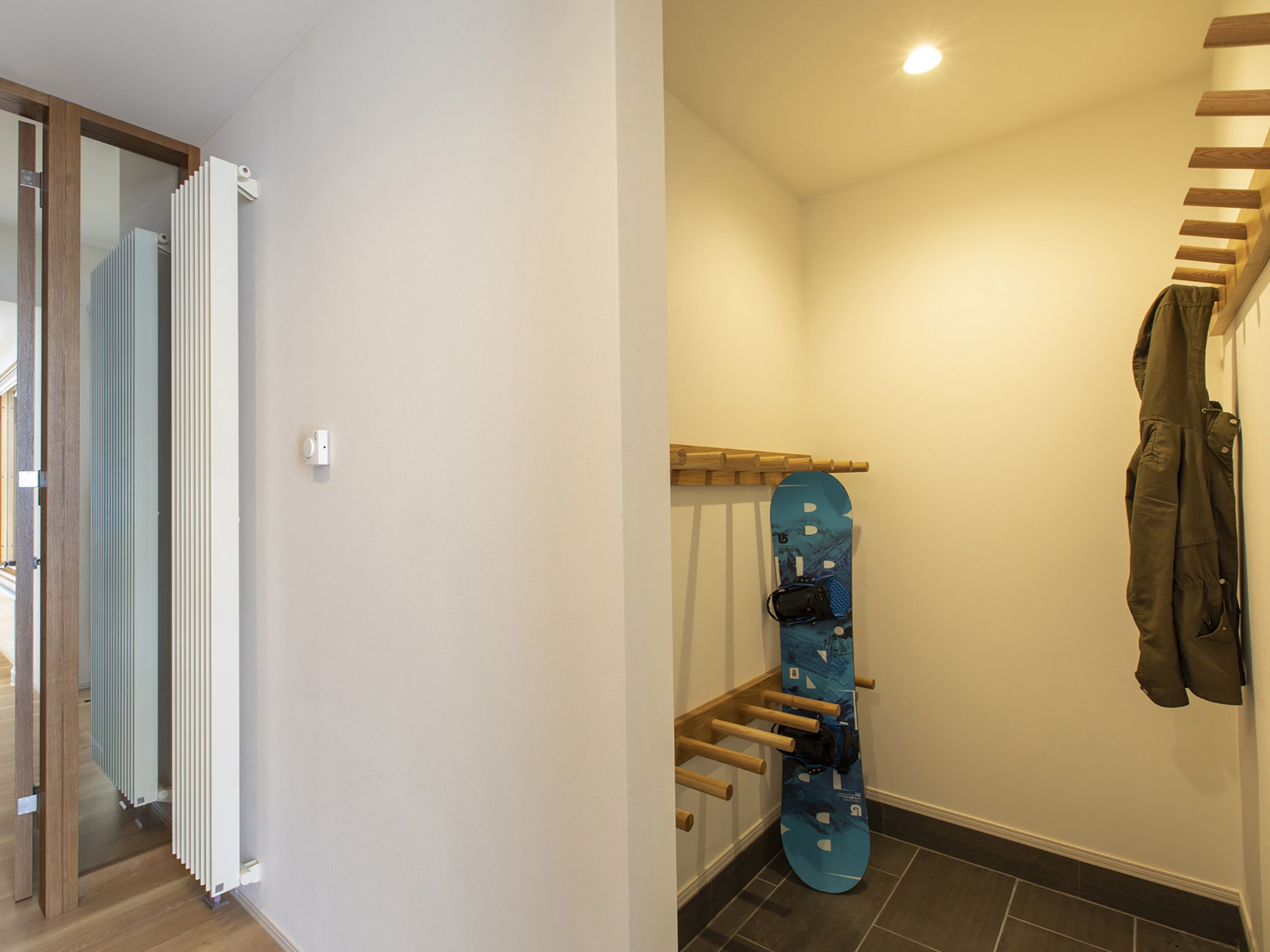 A hallway with a wooden staircase, a blue snowboard leaning against the wall, and a coat hanging on the staircase.