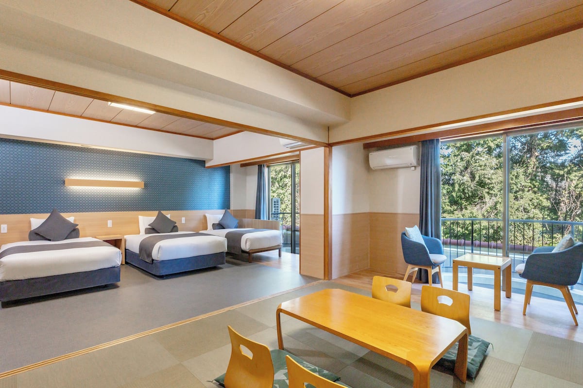 Une chambre d’hôtel moderne dotée d’un grand lit, d’un plus petit lit, d’une table à manger et d’un balcon offrant une vue sur les arbres.