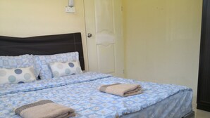 A neatly made bed with a blue floral bedspread and two beige towels, a dark wooden headboard, and a plain wall with a door. - Kuala Mengalong Cottage (Sipitang)