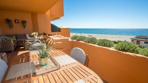 Varanda com mesa de madeira, móveis de vime e um vaso de planta. Tem vista para a praia e para o mar.