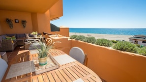 A balcony with a wooden table, wicker furniture, and a potted plant. It overlooks a beach and the sea.