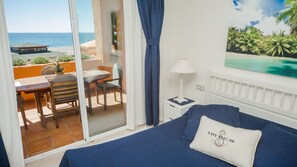 A hotel room with a bed, a nightstand, a lamp, a window with a view of the ocean, and a table with chairs outside.