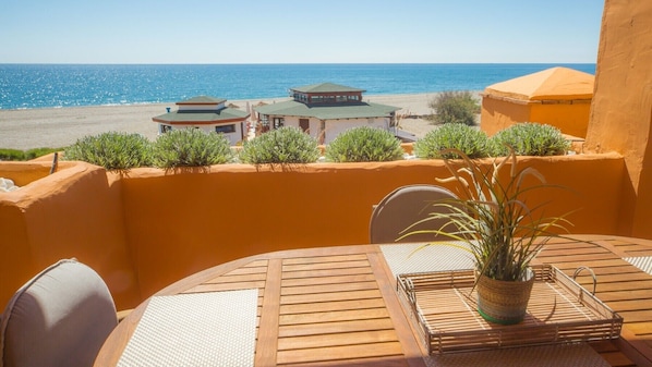 A terrace with a wooden table and chairs, a potted plant, and a view of the beach and ocean.