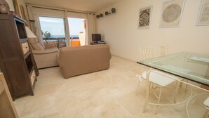 A living room with a sofa, a glass table, and chairs. There is a TV and a shelf with decorative items.