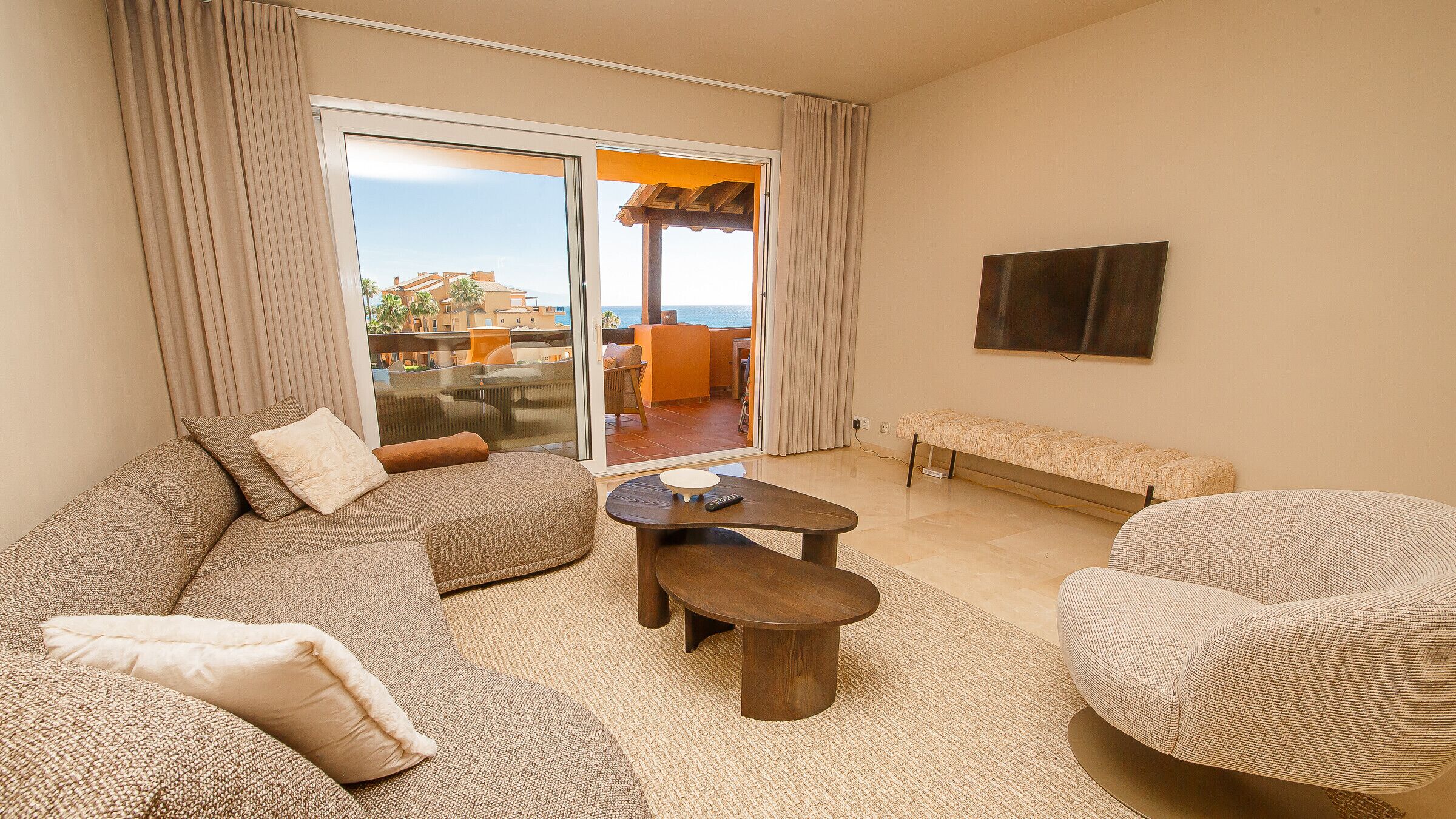 Uma sala de estar com sofá, poltrona, mesa de centro e um banco. Há uma TV fixada na parede e uma porta de vidro deslizante que dá para uma área externa.