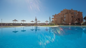 Une piscine aux eaux cristallines, dont la surface reflète l’environnement, entourée de parasols et bordée d’un immeuble à plusieurs étages.
