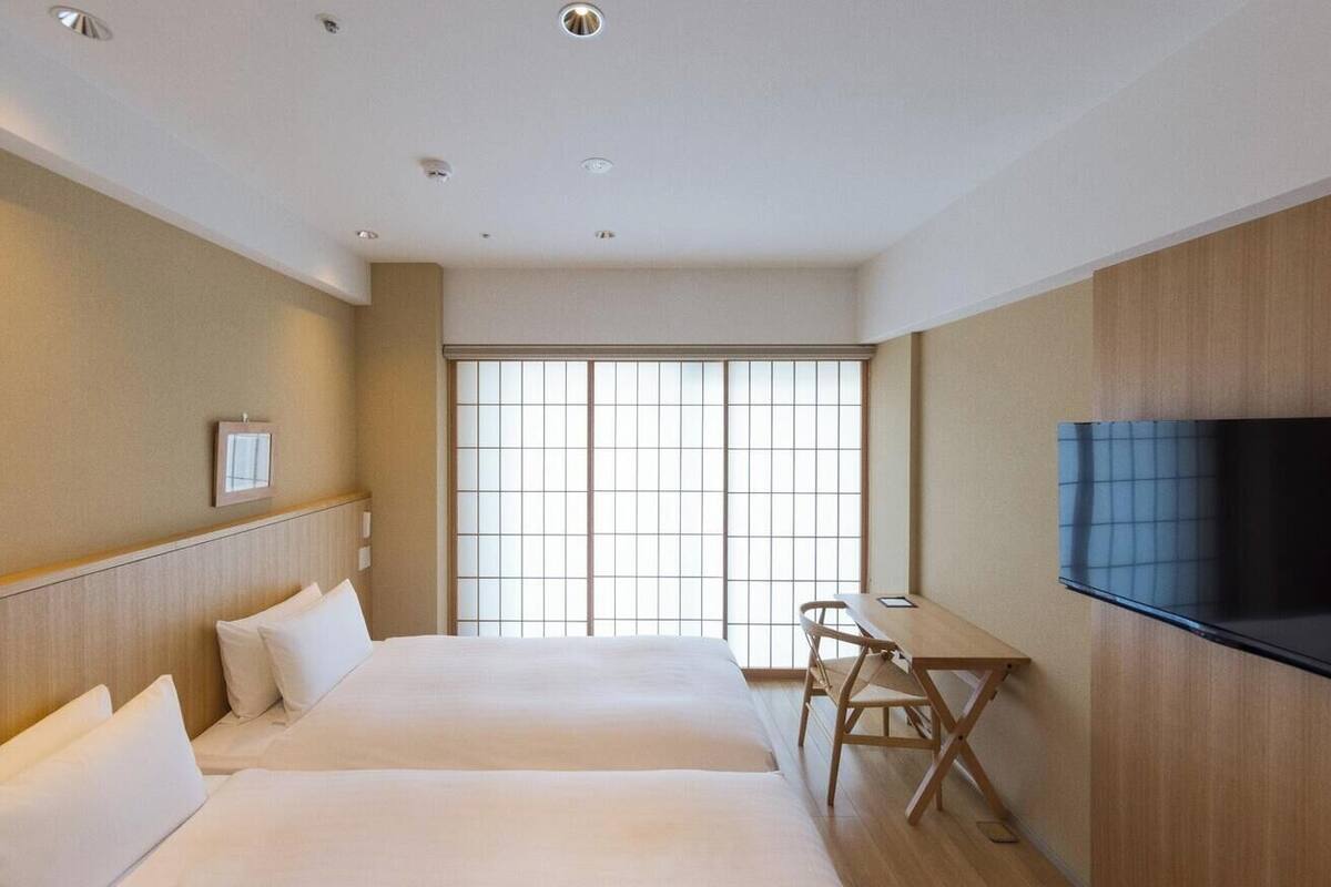 A hotel room with two beds, a desk, a chair, a TV, and a large window.