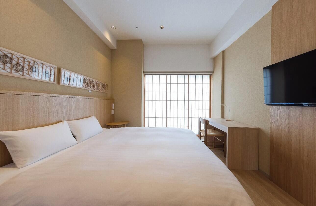 A hotel room with a large bed, a desk, a TV, and a window with blinds.