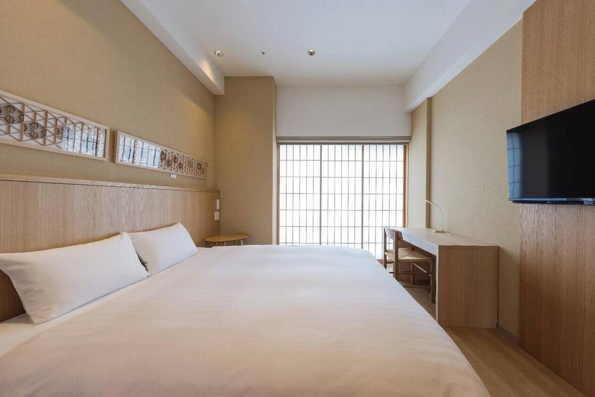 A hotel room with a large bed, a desk, a TV, and a window with blinds.