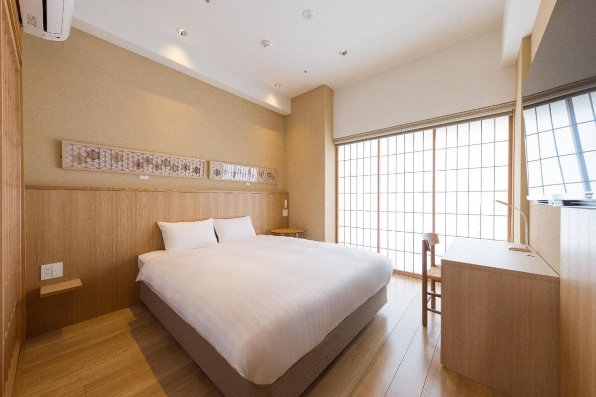 A modern hotel room with a large bed, a desk, and a view of the city through large windows.
