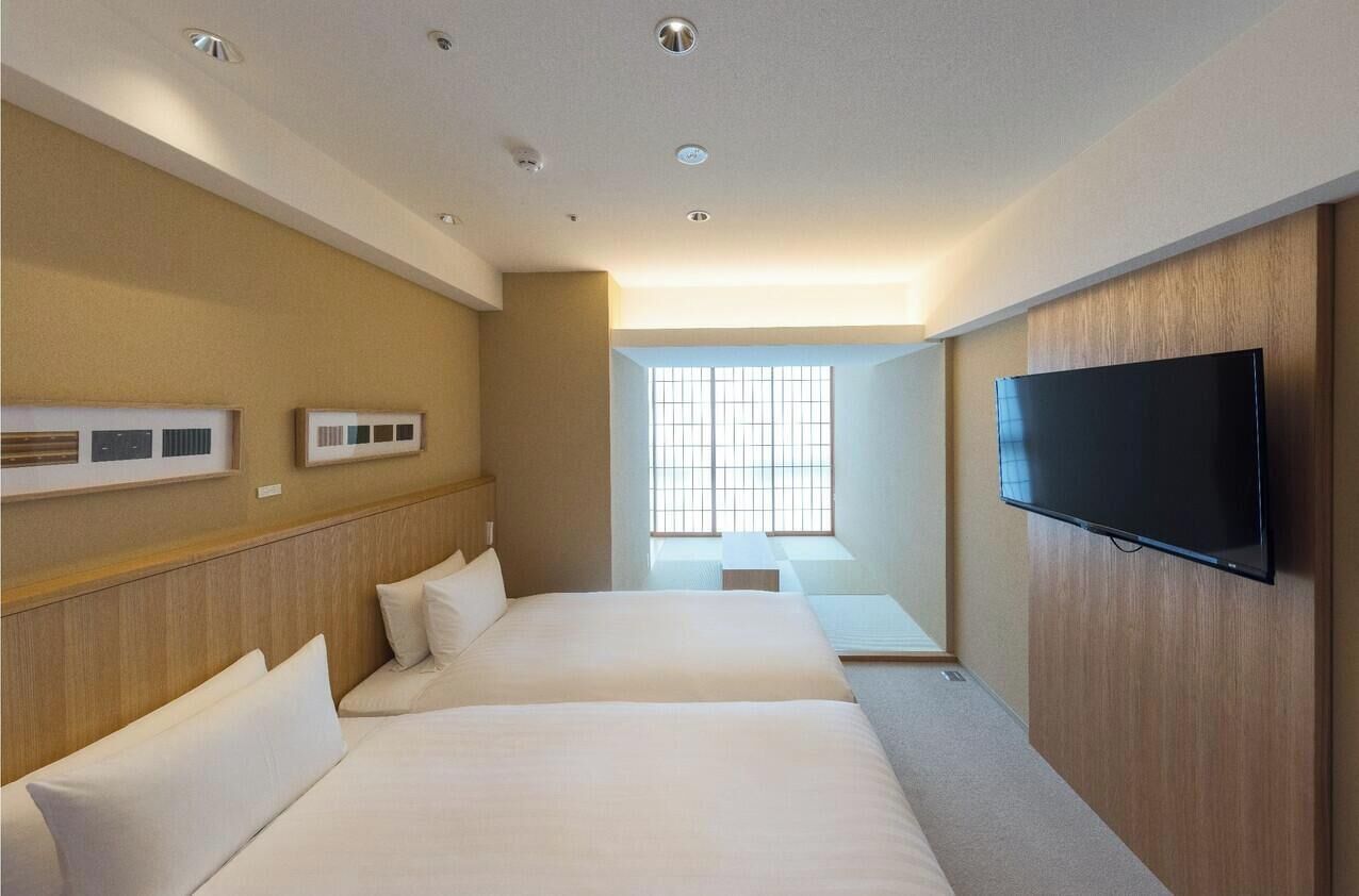 A hotel room with two beds, a flat-screen TV, a window with blinds, and a ceiling with recessed lighting.
