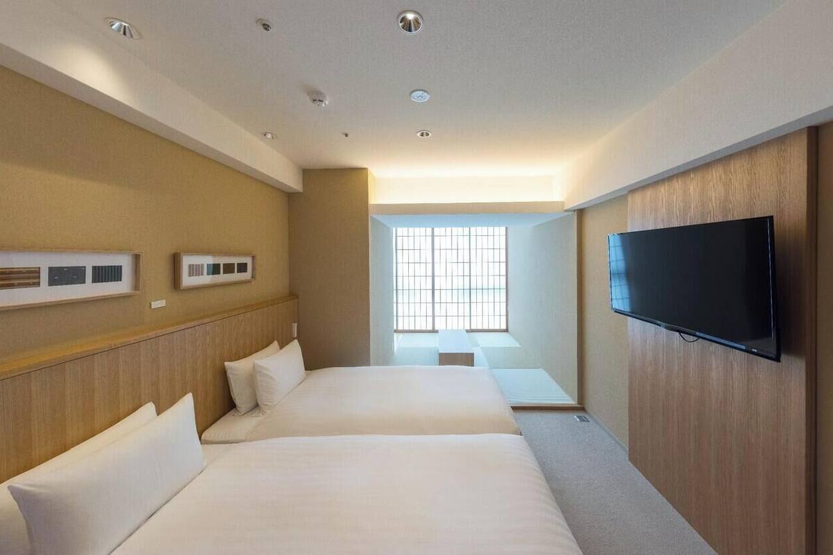 A hotel room with two beds, a flat-screen TV, a window with blinds, and a ceiling with recessed lighting.