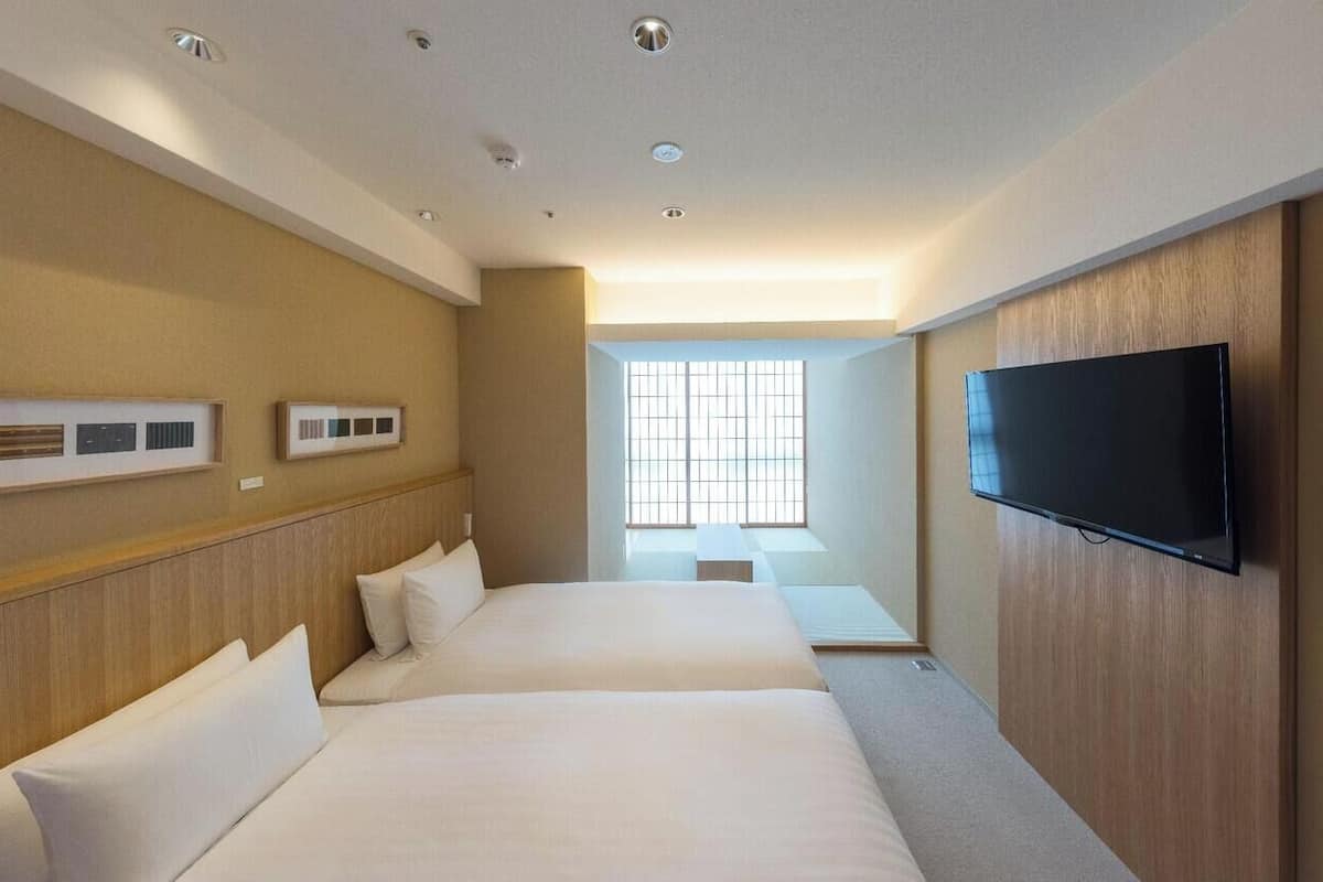 A hotel room with two beds, a flat-screen TV, a window with blinds, and a ceiling with recessed lighting.