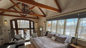 Une chambre spacieuse avec un grand lit, un coin salon comprenant un fauteuil et une table, et une vue sur l’extérieur grâce à de grandes fenêtres.
