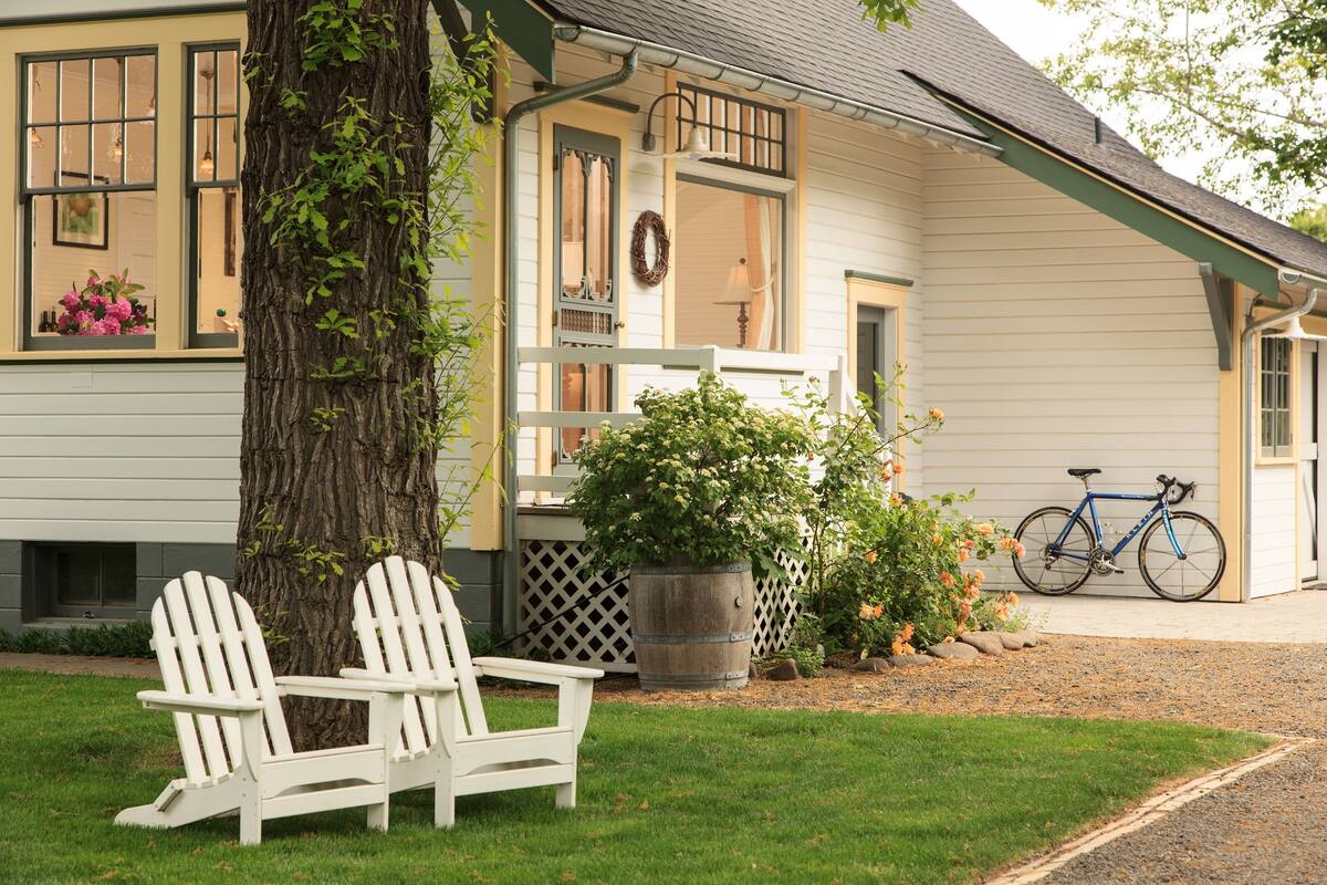 A house with a porch, two Adirondack chairs, a bicycle, and a large tree.