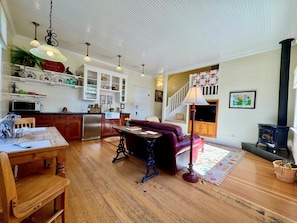 A spacious living area with a wooden floor, a dark red sofa, a dining table with chairs, a kitchen with white cabinets, and a black wood-burning stove.