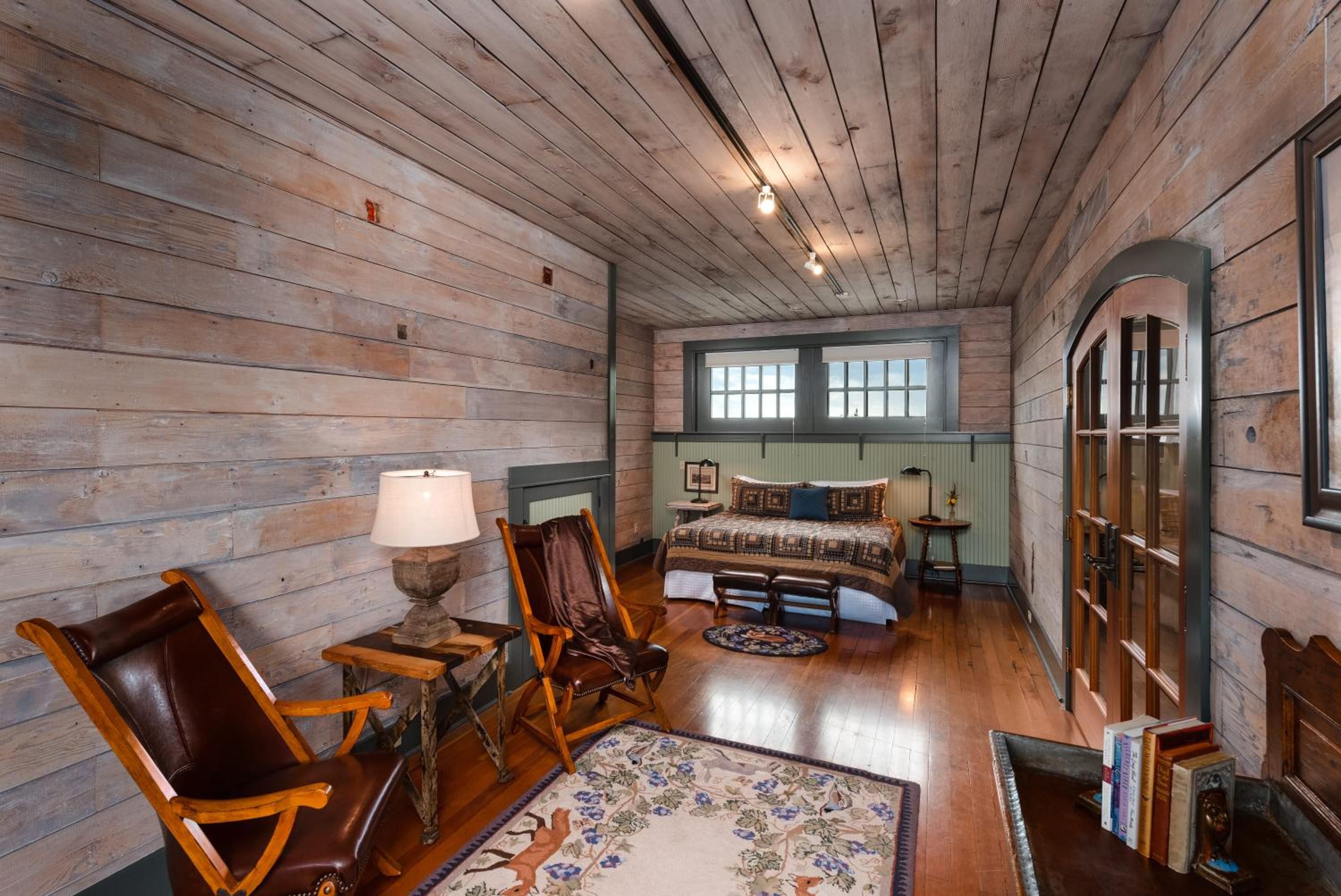 A rustic room with wooden walls, a bed with patterned bedding, a wooden chair, a small table with a lamp, and a window with a view.