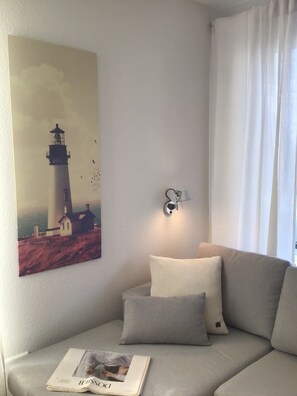 A living room with a beige sofa, a wall-mounted light fixture, and a painting of a lighthouse.