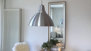 Ein Essbereich mit einem Holztisch, einem weißen Teekessel, Tassen und Untertassen. Eine Topfpflanze, ein flauschiger weißer Sessel und ein Korbsessel sind ebenfalls vorhanden. An der Wand hängt ein großer Spiegel, der den Raum reflektiert.