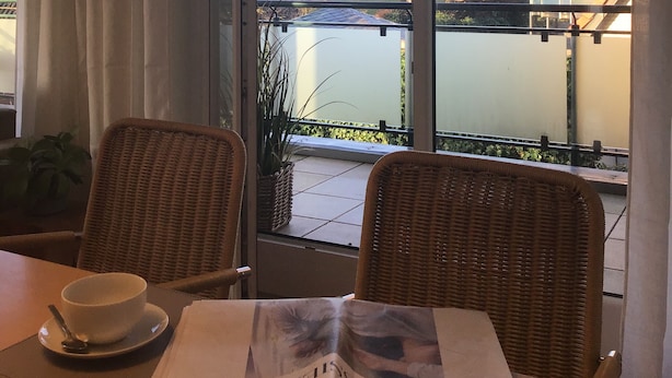 Ein Zimmer mit Blick auf einen Balkon und ein Gebäude gegenüber dem Wasser. Es gibt Korbstühle, einen Tisch mit Tasse und Untertasse sowie eine offene Zeitschrift.