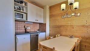 Eine kompakte Küche mit weißen Schränken, einer Mikrowelle, einem Spülbecken und einem Geschirrspüler. Eine Holzvertäfelung zeigt gerahmte Bilder, und ein Esstisch mit Stühlen lädt zum Verweilen ein.