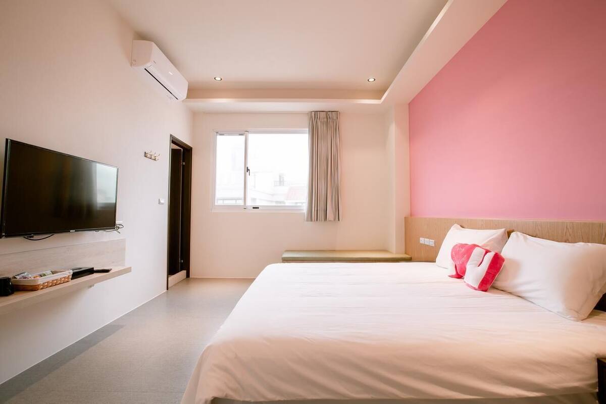 A hotel room with a large bed, a flat-screen TV mounted on the wall, a window with curtains, and a pink accent wall.