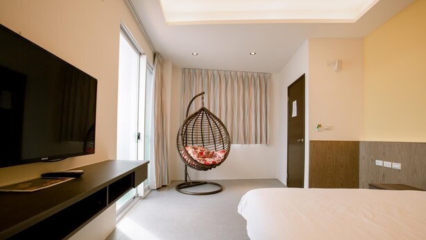 Ein modernes Hotelzimmer mit einem großen Bett, einem Fernseher auf einer dunklen Kommode, einem Hängekorbsessel mit Blumenkissen und einem großen Fenster mit Vorhängen.
