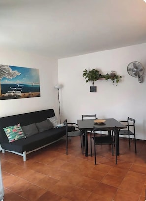A living room with a black sofa, a dining table with chairs, a painting on the wall, a floor lamp, and a wall-mounted fan.