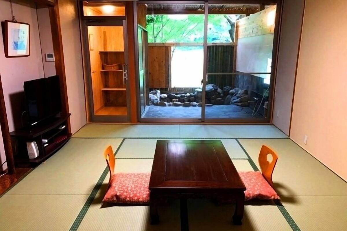 A tatami mat room with a wooden table and chairs, a television, and a sliding door leading to a garden.