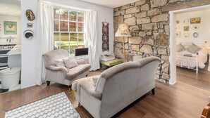 A cozy living room with two grey sofas, a stone wall, a kitchen area with a stove, and a dining table with chairs.