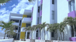 A modern building with white and purple facades, multiple balconies, and a paved walkway with palm trees. - AMA Yucatan (Progreso)