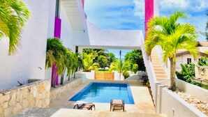 A modern pool area with a clear view of the ocean, surrounded by palm trees and a white building with pink accents. - AMA Yucatan (Progreso)