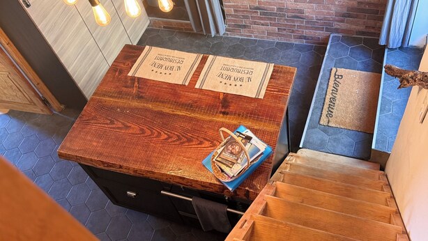 A wooden table with a placemat, a basket, and a magazine. There are hanging light bulbs, a brick wall, and a wooden staircase. - Atypical village apartment (Olmeto)