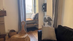 A cozy living room with a black sofa, a wicker chair, a fan, and a window with curtains. - Atypical village apartment (Olmeto)