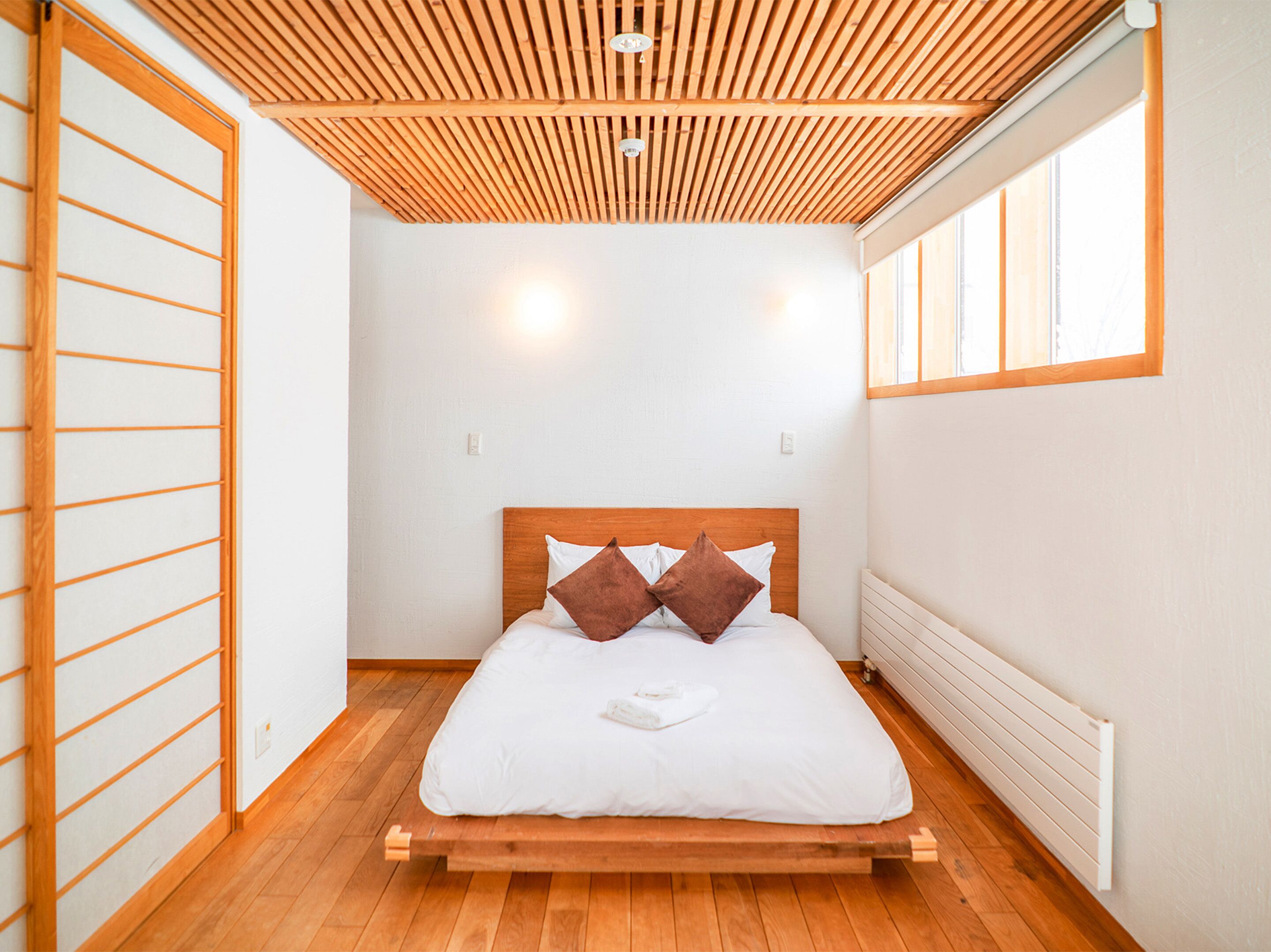 A minimalist bedroom with a wooden bed frame, white bedding, and a slatted wooden ceiling.