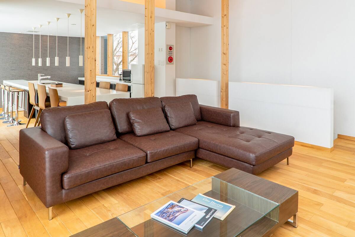 Ein modernes Wohnzimmer mit einem braunen Ledersofa, einem Glastisch und einer Küchenzeile im Hintergrund.