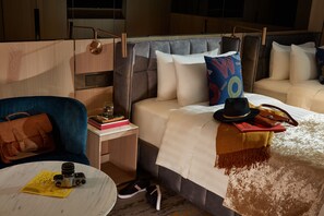 Une chambre d’hôtel moderne avec un lit, un fauteuil bleu, une petite table sur laquelle se trouve un appareil photo, et une table de chevet avec des livres et une lampe.