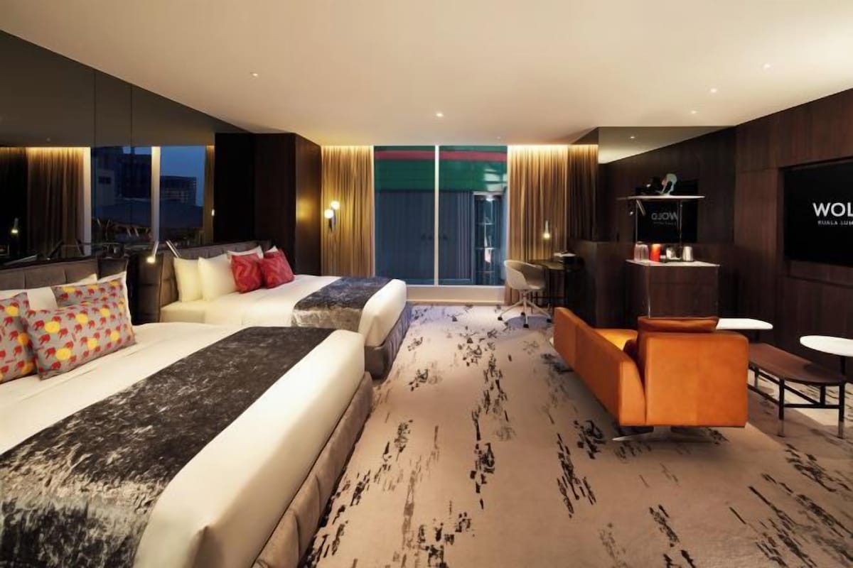 A modern hotel room with a large bed, a sofa, an orange armchair, a small table, and a TV mounted on the wall.