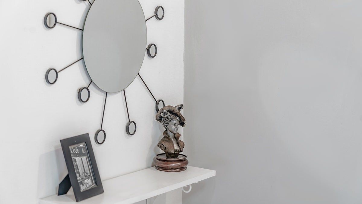 A white wall with a circular mirror surrounded by rings, a bust sculpture, and a framed picture on a white shelf.