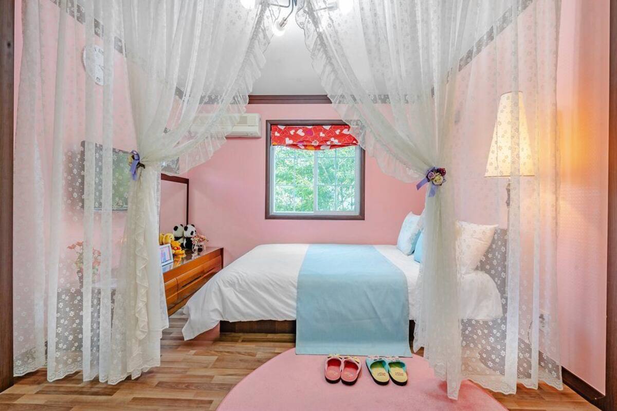 Un dormitorio con cama, mesita de noche, lámpara, ventana y un otoman rosa con zapatillas.